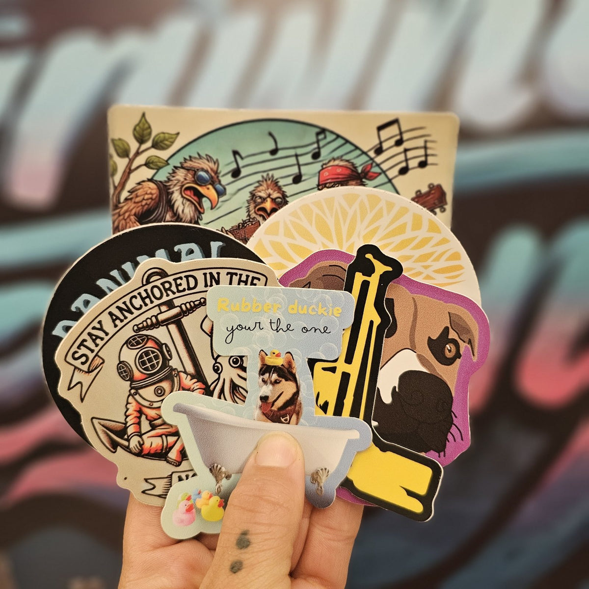 Hand holding a stack of custom stickers in front of the Fairwinds Designs mural
