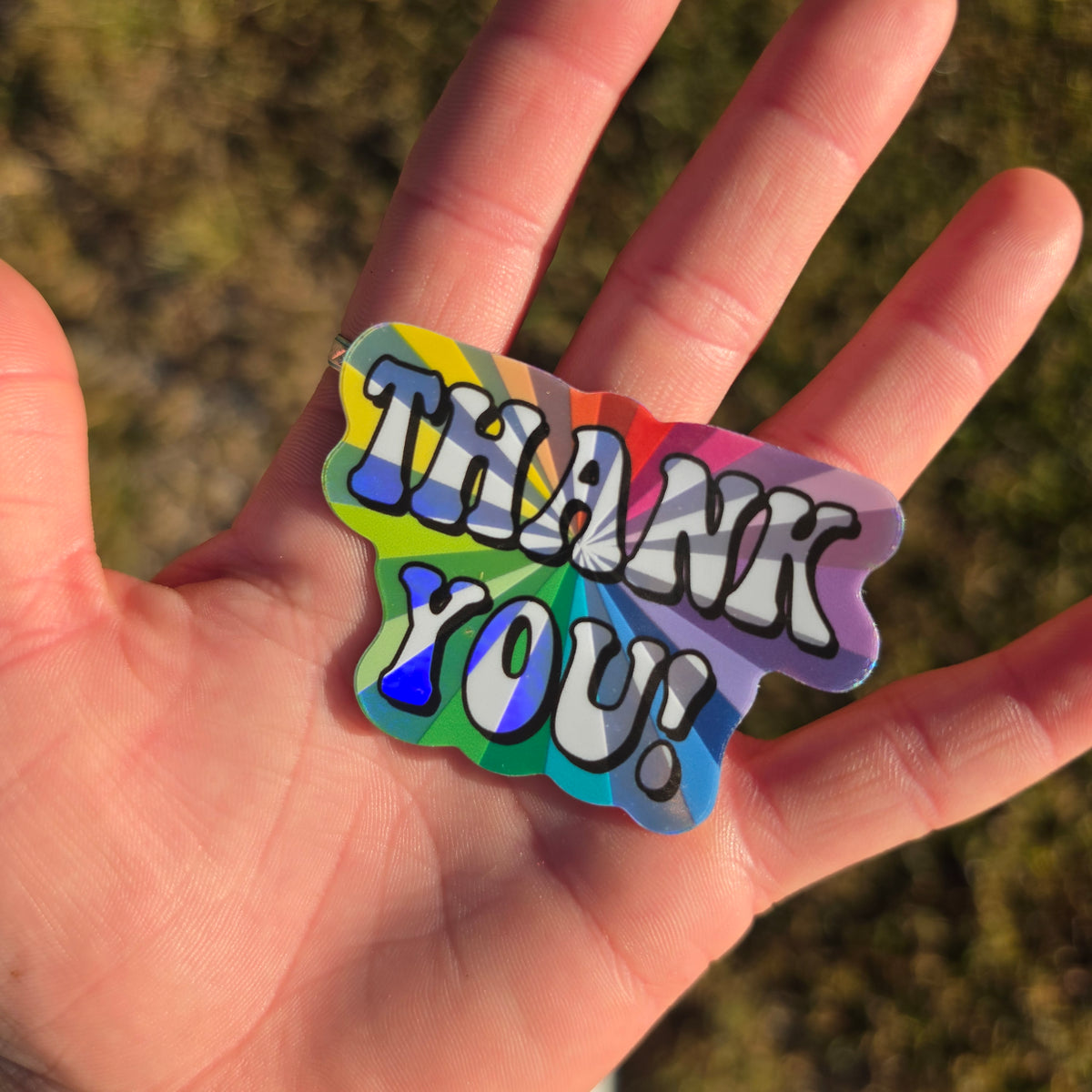 Holographic Thank You! sticker with a rainbow sunburst design, held in an open hand