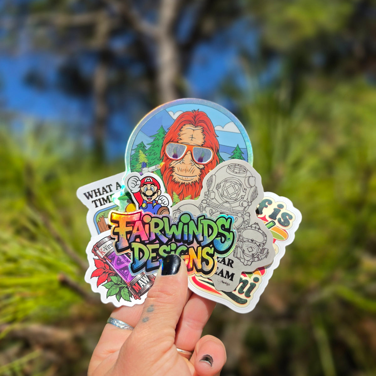 Hand holding a mix of holographic and printed stickers, including a Fairwinds Designs logo sticker