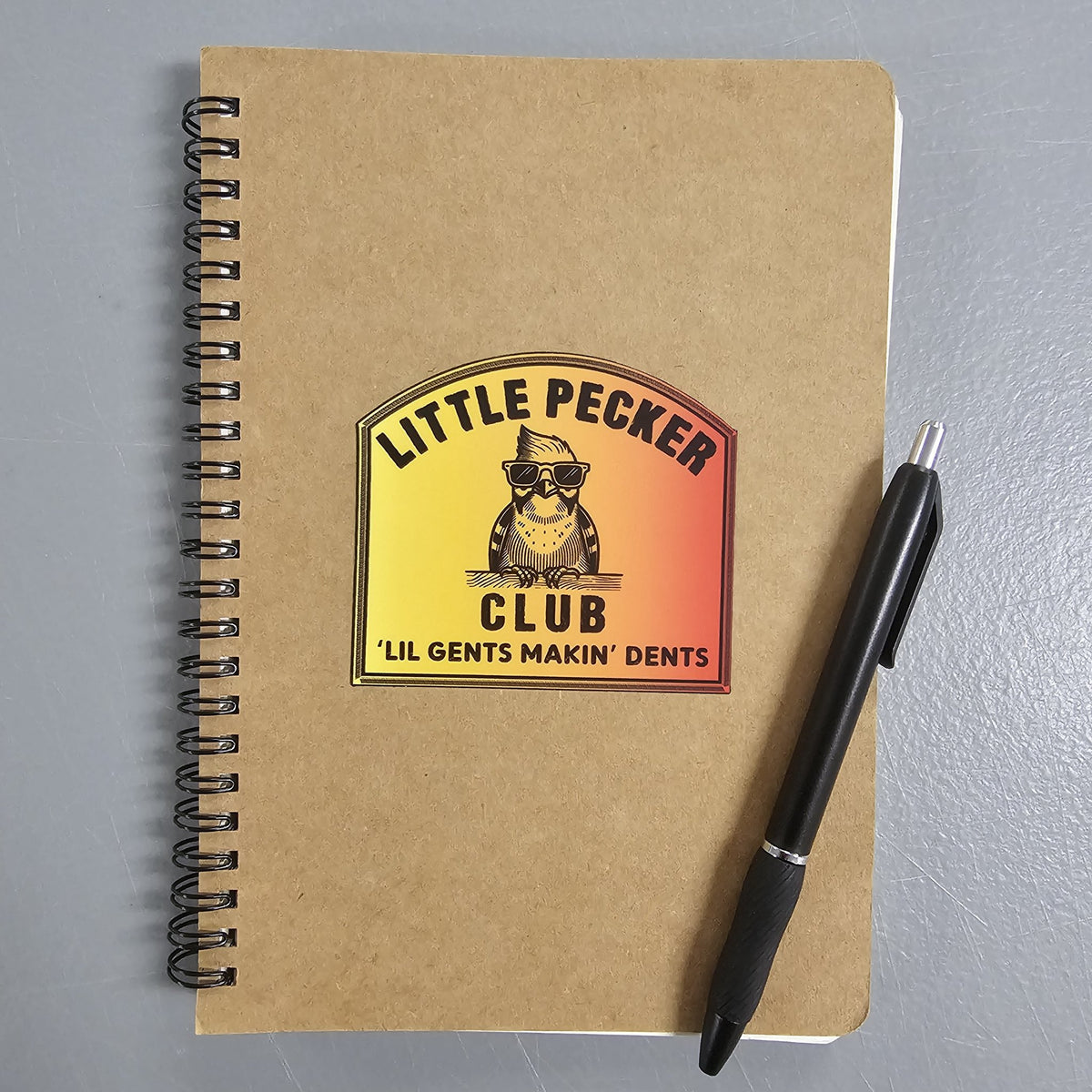 Little Pecker Club sticker on a kraft paper notebook next to a pen